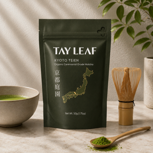 A stand-up pouch of Tay Leaf Kyoto Teien organic ceremonial grade matcha stands on a light stone surface, with a bowl of green tea, a bamboo whisk, and a wooden spoon of matcha powder nearby. A leafy plant and textured wall are softly blurred in the background.